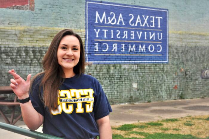 Picture of Heather Rodriguez making the lion sign with behind a mural of Texas A&M University Commerce.